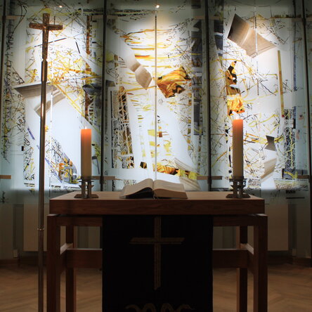 Ein Andachtsraum mit gold-weißen Farbakzenten. Auf einem Altar brennen zwei Kerzen. Daneben steht ein Kreuz.